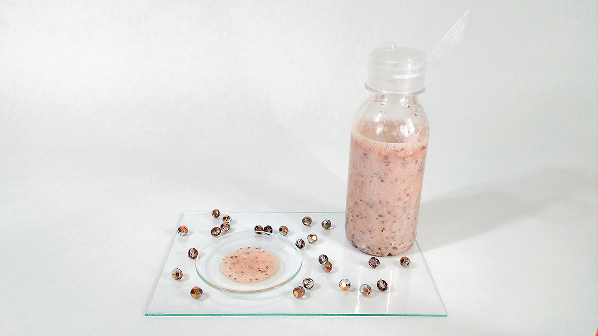 Peelinogvý sprchový gél ružovej farby s jahodovými zrnkami vo fľaške a kvapka na priehľadnej sklenej podložke na bielom pozadí.